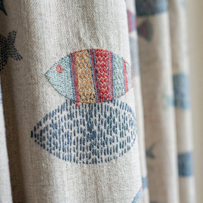 May include: Beige fabric curtain with embroidered fish and starfish designs. The fish are in shades of blue, red, and yellow, with a detailed stitched pattern. The starfish and other shapes are in blue. The fabric has a textured appearance.
