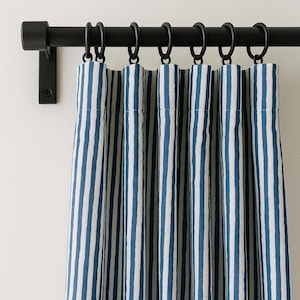 Può includere: Un bastone per tende nero con anelli abbinati sostiene un set di tende a righe blu e bianche. Le tende sono appese contro un muro bianco, creando un aspetto pulito e classico. Il bastone è montato con una staffa nera.