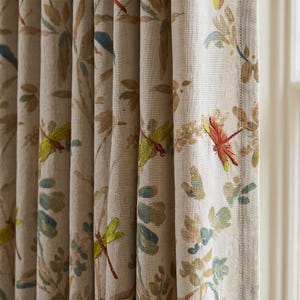 Op de afbeelding: Een beige gordijn met een herhalende botanische print met geborduurde libellen in tinten oranje, geel en groen. De stof heeft een zichtbare geweven textuur en het gordijn is in verticale plooien langs de bovenrand verzameld.
