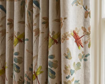 May include: A beige curtain featuring a repeating botanical print with embroidered dragonflies in shades of orange, yellow, and green. The fabric has a visible woven texture, and the curtain is gathered into vertical pleats along the top edge.