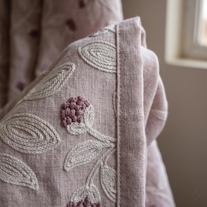Könnte beinhalten: Ein altrosa Leinenstoff mit aufwendiger Stickerei aus cremefarbenen Blättern und dunkelmalvenfarbenen Blütenknospen. Das Textil weist eine sichtbare Webstruktur mit einem sauber vernähten Saum entlang der Kante auf und zeigt ein detailliertes botanisches Muster.