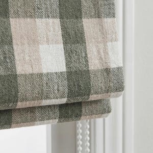 May include: Close-up of a green, beige, and white checkered fabric roman shade. The fabric is textured and appears to be linen or a similar material. The shade is partially rolled up, revealing the layered construction and a white beaded pull cord.