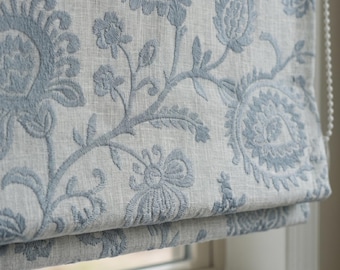 May include: A roman blind featuring a light grey fabric base with an intricate, raised embroidery pattern in a dusty blue colour. The design consists of stylised floral and paisley motifs that repeat across the surface, paired with a white beaded chain.