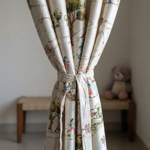 May include: Cream-colored curtain with a whimsical print featuring trees, figures, and a road. The curtain is tied with a matching fabric tie. A bench and a teddy bear are visible in the background.