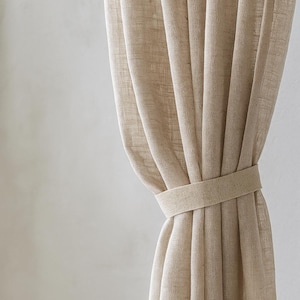 Puede incluir: Cortinas de lino beige con una cinta a juego. Las cortinas están hechas de una tela texturizada y están parcialmente corridas, revelando los pliegues y el diseño simple de la cinta. La paleta de colores neutros sugiere una estética natural y minimalista.