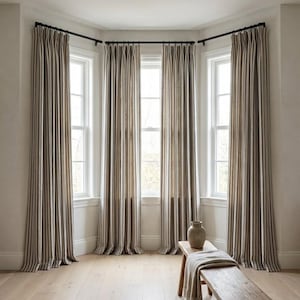 Op de afbeelding: Een kamer met drie ramen, elk omlijst door lange, verticaal gestreepte gordijnen in bruin, beige en zwart. De gordijnen hangen aan zwarte stangen. Een houten bank met een vaas en een opgevouwen deken staat voor de ramen.