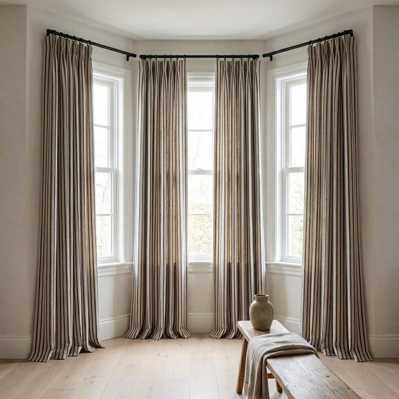 Op de afbeelding: Een kamer met drie ramen, elk omlijst door lange, verticaal gestreepte gordijnen in bruin, beige en zwart. De gordijnen hangen aan zwarte stangen. Een houten bank met een vaas en een opgevouwen deken staat voor de ramen.