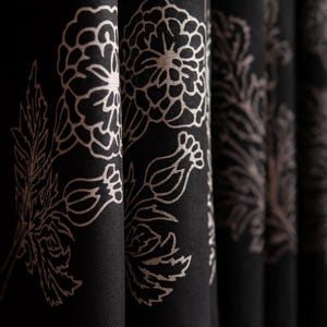 Puede incluir: Primer plano de cortinas de tela negra con un estampado floral. El diseño presenta grandes flores estilizadas y hojas en color beige claro. Las cortinas están dobladas, creando un efecto texturizado y en capas.