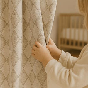 May include: Close-up of a beige curtain with a diamond pattern. The curtain is being held open by a person. The curtain has a textured appearance and is a neutral color. The background includes a crib.