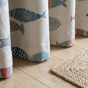 May include: Close-up of beige fabric curtains with a repeating pattern of colorful fish in blue, red, and orange. The curtains are hanging in front of a light brown wooden floor, with a small, woven jute rug in the foreground.