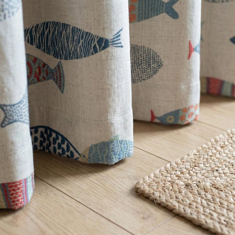 May include: Close-up of beige fabric curtains with a repeating pattern of colorful fish in blue, red, and orange. The curtains are hanging in front of a light brown wooden floor, with a small, woven jute rug in the foreground.