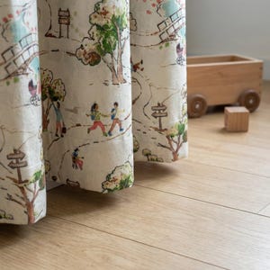 May include: Cream-colored curtains with a whimsical illustrated design of a park scene. The fabric features a path, trees, bridges, and figures of people. A wooden toy wagon and block sit on the light wood floor.