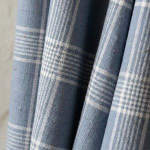 May include: Close-up of blue and white plaid fabric. The fabric has a woven texture with a classic plaid pattern. The blue color is a soft, muted shade, and the white lines create a grid-like design. The fabric is draped, showing folds and shadows.