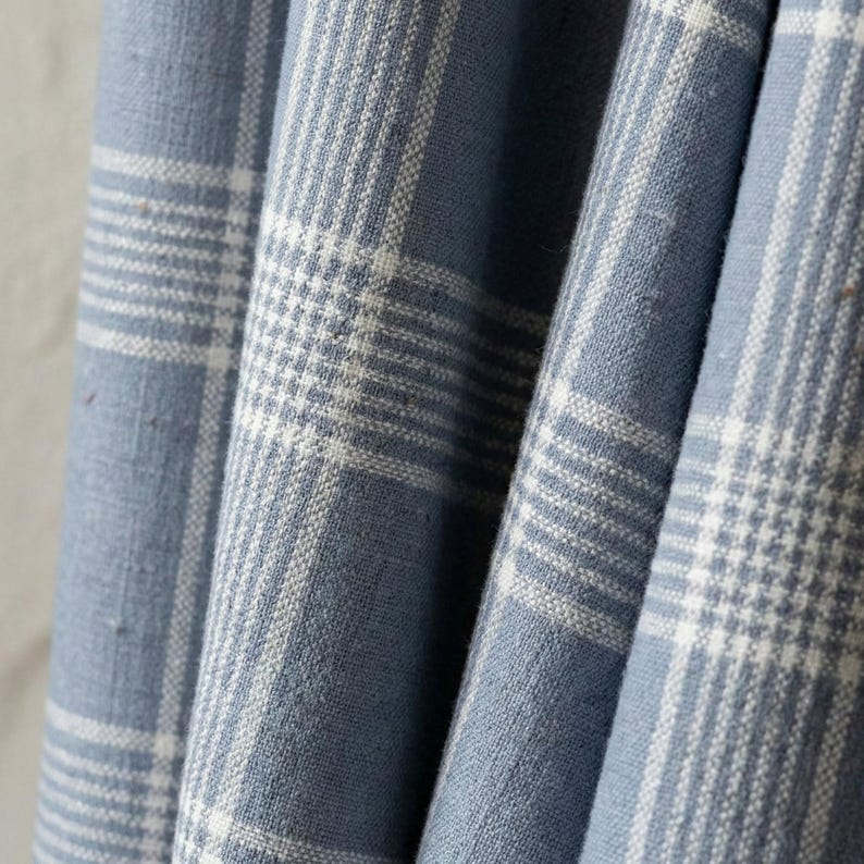 May include: Close-up of blue and white plaid fabric. The fabric has a woven texture with a classic plaid pattern. The blue color is a soft, muted shade, and the white lines create a grid-like design. The fabric is draped, showing folds and shadows.