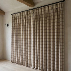 May include: Beige and brown plaid curtains hanging from a black rod. The curtains are pleated and cover a large window. The room has light-colored walls and a wooden beam.