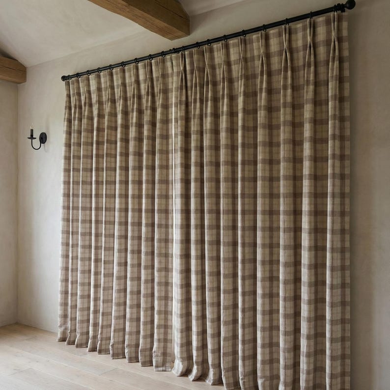 May include: Beige and brown plaid curtains hanging from a black rod. The curtains are pleated and cover a large window. The room has light-colored walls and a wooden beam.