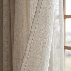 Puede incluir: Primer plano de cortinas de lino beige. La tela tiene un aspecto texturizado con hilos tejidos visibles. Las cortinas están parcialmente abiertas, revelando un vistazo de una ventana y la vista exterior. El color neutro y la textura natural sugieren un estilo rústico o minimalista.