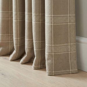 May include: Close-up of beige plaid curtains. The curtains are made of a thick fabric with a grid pattern in a lighter beige color. The curtains are hanging in front of a light gray wall and a wooden floor.