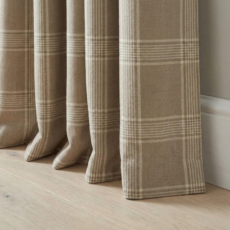 May include: Close-up of beige plaid curtains. The curtains are made of a thick fabric with a grid pattern in a lighter beige color. The curtains are hanging in front of a light gray wall and a wooden floor.