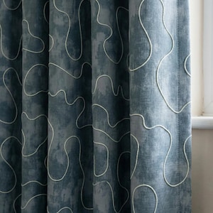 May include: Close-up of a blue curtain with a textured, mottled appearance. White, abstract, wavy lines are embroidered across the fabric. The curtain is hanging in front of a window, with the edge of the window frame visible.