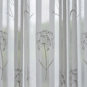 K&ouml;nnte beinhalten: Ein halbtransparenter wei&szlig;er Vorhang mit einem sich wiederholenden vertikalen Muster aus gestickten grauen botanischen Bl&uuml;tenk&ouml;pfen und St&auml;ngeln, die an L&ouml;wenzahn erinnern. Der Stoff ist leicht und durchscheinend und l&auml;sst Licht hindurch.
