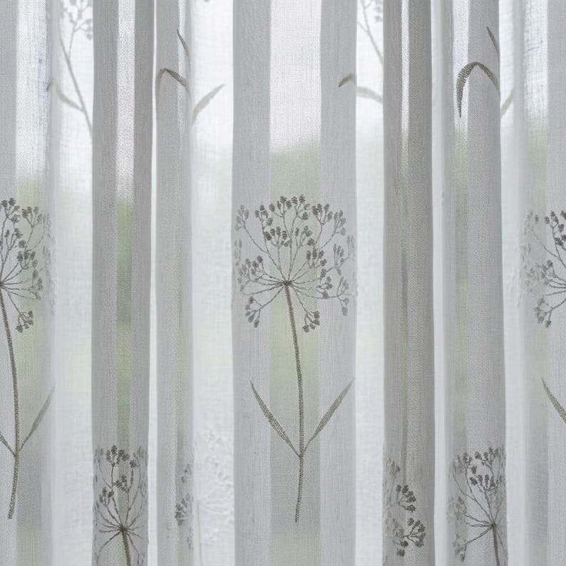 K&ouml;nnte beinhalten: Ein halbtransparenter wei&szlig;er Vorhang mit einem sich wiederholenden vertikalen Muster aus gestickten grauen botanischen Bl&uuml;tenk&ouml;pfen und St&auml;ngeln, die an L&ouml;wenzahn erinnern. Der Stoff ist leicht und durchscheinend und l&auml;sst Licht hindurch.