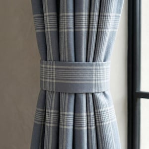 May include: Blue plaid curtains with a matching fabric tie-back. The curtains are a light blue color with a white plaid pattern. The tie-back is a solid light blue color with a white plaid pattern.