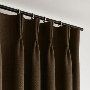 May include: Close-up of brown herringbone patterned curtains hanging from a black rod with silver rings. The curtains are pleated and create vertical folds. The fabric has a subtle texture and a warm, neutral color.