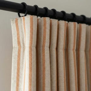 May include: Close-up of a set of striped curtains in beige and orange hues. The curtains are hanging from a black metal rod with black rings. The fabric has vertical stripes, creating a simple, yet elegant design.