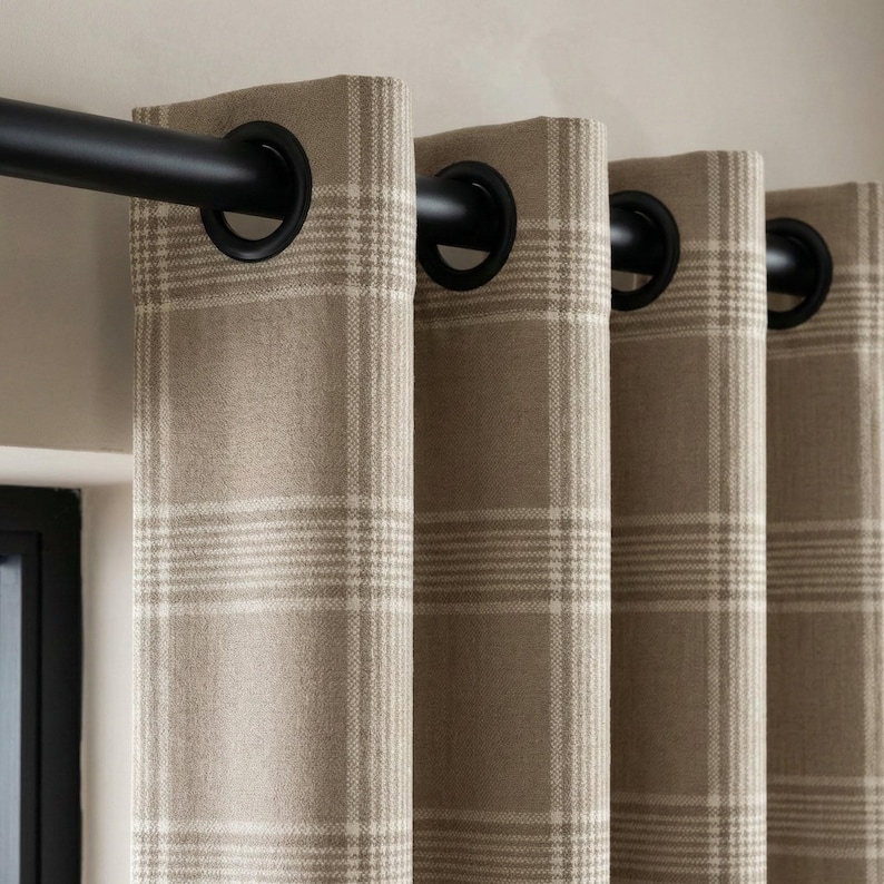 May include: Close-up of beige plaid curtains with black grommets and a black curtain rod. The curtains have a neutral color palette with a grid pattern. The window treatment is a home decor item.