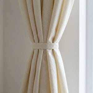 Può includere: Tende in lino color crema, legate con una fascia in tessuto abbinata. Le tende sono drappeggiate, mostrando morbide pieghe e una texture naturale. La palette di colori neutri crea un'atmosfera calma ed elegante.
