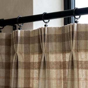 May include: Close-up of a window treatment featuring a black curtain rod with rings and pleated, beige and brown plaid curtains. The curtains are made of a textured fabric and hang in soft folds, creating a classic, neutral aesthetic.