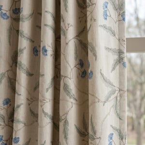 Könnte beinhalten: Ein beigefarbener Vorhang mit einem gestickten Muster aus blauen Blumen und grünen farnartigen Blättern. Der Stoff hat eine sichtbare Webstruktur und das Design wiederholt sich über die vertikalen Falten des Vorhangs.