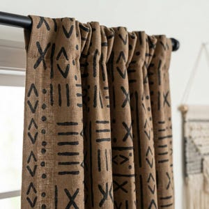 May include: Brown curtains with a repeating black geometric pattern. The curtains are hanging from a black rod. The pattern includes shapes like chevrons, lines, and dots. The fabric appears to be textured.