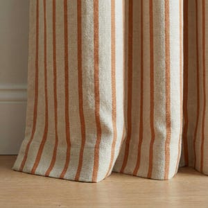 May include: Close-up of beige and orange striped curtains. The vertical stripes are a warm orange color against a neutral beige background. The curtains are made of a textured fabric and are partially open, revealing a glimpse of the floor.