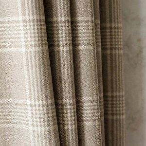 May include: Close-up of a beige and white plaid fabric, likely a curtain or drapery. The fabric is folded, showing the texture and pattern of the woven material. The plaid design features a grid of intersecting lines, creating a classic and neutral aesthetic.