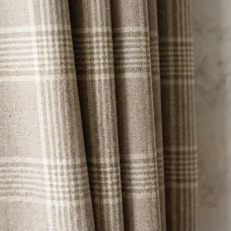 May include: Close-up of a beige and white plaid fabric, likely a curtain or drapery. The fabric is folded, showing the texture and pattern of the woven material. The plaid design features a grid of intersecting lines, creating a classic and neutral aesthetic.