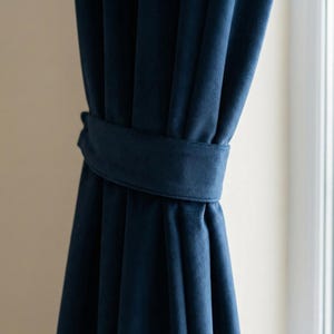 Puede incluir: Primer plano de cortinas de terciopelo azul marino, sujetas por un recogedor de tela a juego. Las cortinas están drapeadas, con pliegues suaves y una textura lujosa. La luz natural entra por una ventana en el fondo.