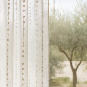 May include: Off-white linen curtain with vertical rows of small, tan, circular embellishments. The curtain hangs in front of a blurred outdoor scene with a tree. The curtain's texture is visible, suggesting a light and airy fabric.