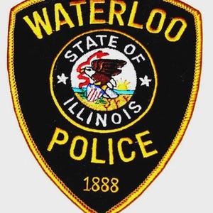 May include: A black and gold police patch with the words "WATERLOO POLICE" and "1888". The patch features a circular emblem with an eagle, the words "STATE OF ILLINOIS", and a flag.