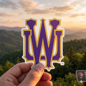 May include: A sticker featuring a purple and gold stylized "W" logo with a white border. The sticker is held up against a backdrop of a mountain range and a sunset.