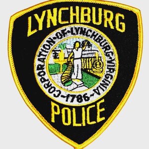 May include: A shield-shaped patch with a black background and a gold border. The patch features the text "LYNCHBURG POLICE" in yellow, with a central emblem depicting a figure and the text "CORPORATION OF LYNCHBURG VIRGINIA 1786".
