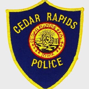 May include: A blue and yellow police patch with the words "CEDAR RAPIDS POLICE" in yellow. The center features a circular emblem with the words "CEDAR RAPIDS SEAL IOWA". The patch has a shield shape with a yellow border.