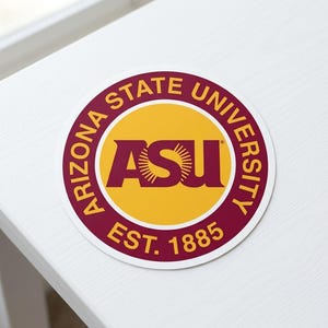 May include: A round sticker featuring the Arizona State University logo. The logo includes the letters "ASU" in maroon, set against a yellow circle. The words "ARIZONA STATE UNIVERSITY" and "EST. 1885" are in maroon around the circle's edge.