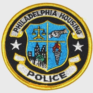 May include: Embroidered patch featuring the words "PHILADELPHIA HOUSING POLICE" in a circular design. The patch has a black background with a blue shield in the center, displaying a scale, a handshake, a cityscape, and a torch. Two white stars flank the shield.