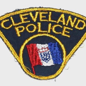 May include: Embroidered patch featuring the words "CLEVELAND POLICE" in gold thread on a navy blue background. A circular emblem in the center displays a flag with red, white, and blue stripes and a crown.
