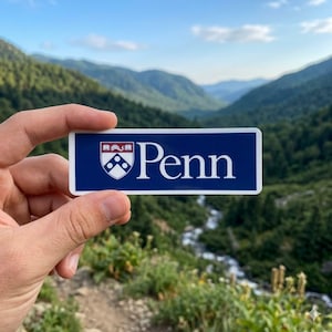May include: A rectangular sticker with a white border and a dark blue background. The sticker features the word "Penn" in white, along with a crest. The crest includes a shield and a banner. The sticker is held up against a scenic mountain backdrop.