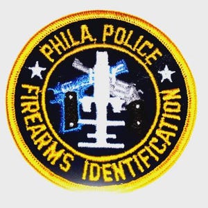 May include: Circular patch with yellow embroidered border. The patch features the text "PHILA. POLICE FIREARMS IDENTIFICATION" in yellow. A white emblem is in the center, with blue and silver gun imagery. Two white stars are on the patch.