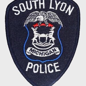 May include: A navy blue police patch with white lettering that reads "SOUTH LYON" at the top and "POLICE" at the bottom. The patch features an eagle and the words "MICHIGAN" in a banner.