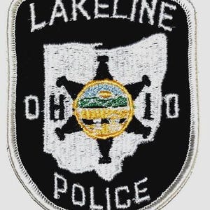May include: A black and white Lakeline Police patch. The patch features the text "LAKELINE" at the top, "POLICE" at the bottom, and "OHIO" on either side of a central emblem. The emblem is a gold star with a blue, green, and yellow circular design in the center.